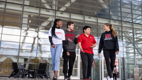 A group of four students walking and talking in front of a building with a glass facade.